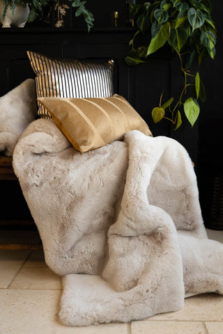 The Natural Faux Fur Throw draped across a bench with some cushions on, with various greenery in the background.