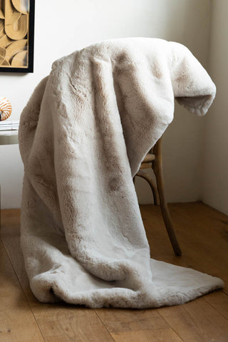 The Natural Faux Fur Throw styled draped over a chair in the corner of a room, with a table, ornament and framed artwork.