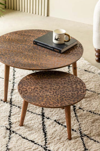 The Natural Leopard Love Nest Of Coffee Tables styled with a book and mug on, displayed on a rug next to a chair, with a plant in the background.