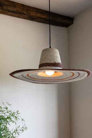 The Natural Seagrass Ceiling Light switched on and hanging in front of a neutral wall with a plant.