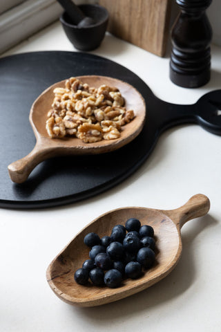 The Natural Wood Carved Serving Dish with the round version, displayed together in a kitchen, styled with nuts, berries and various kitchen accessories.