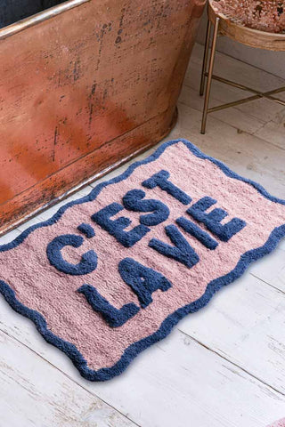 The Navy & Pale Pink C'est La Vie Bath Mat displayed on a bathroom floor next to a bath and side table.