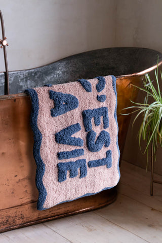 The Navy & Pale Pink C'est La Vie Bath Mat draped across the edge of a copper bath, styled with a plant.