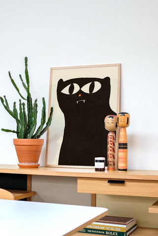 A stylish Noir Cool Cat art print featuring a black cat with large, wide eyes and tiny fangs, displayed unframed on a wooden desk. The setup includes a potted cactus and two traditional wooden Japanese Kokeshi dolls.