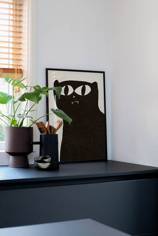 Modern art print of a black cat with a minimalist design, featuring big, bold eyes and a small fang, displayed on a black chest of drawers.