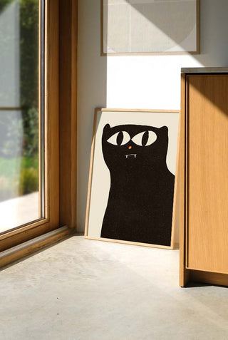 nframed art print of a black cat with oversized eyes and a small orange nose, resting on the floor and leaning against the wall.