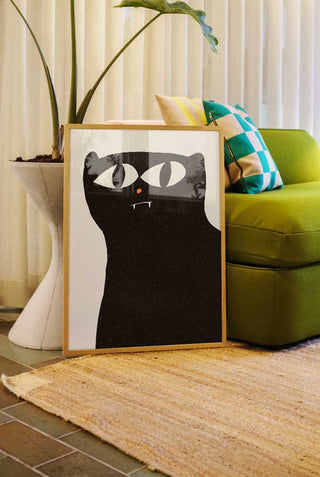 A contemporary art print of a black cat with exaggerated eyes and small fangs in a living room. The print is framed leaning against a green sofa.