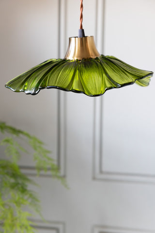 The Olive Green Flower Glass Easyfit Ceiling Light Shade hanging in front of a white wall with a plant.