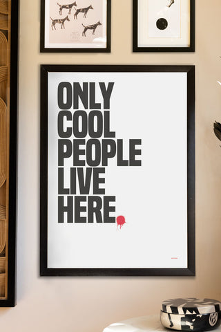 The Only Cool People Live Here Art Print - Unframed styled in a black frame on a neutral wall with some other art prints, above a table with a monochrome trinket box.