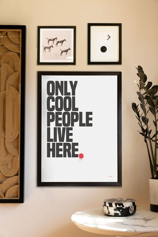 The Only Cool People Live Here Art Print - Unframed styled in a black frame on a neutral wall with other art prints, above a round marble table styled with a trinket box and a potted plant.