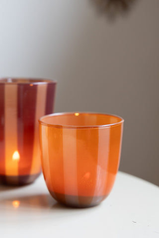 The Orange & Pink Tones Striped Tealight Holder - Short with a lit candle inside, displayed on a table with another version in the background.