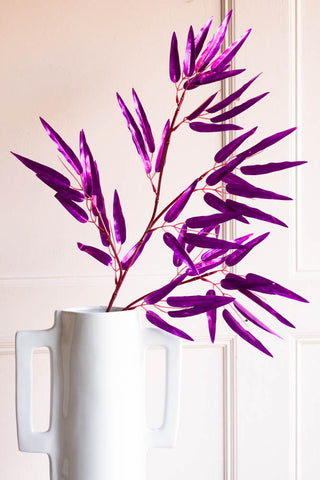 The Ornamental Bamboo In Purple displayed in a white vase in front of a plain wall.