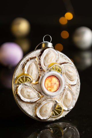 The Oyster Christmas Tree Decoration styled on a black surface with baubles and fairy lights in the background.