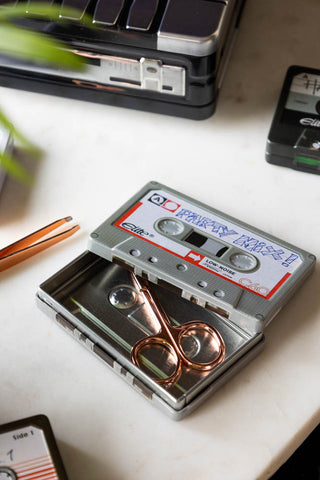 The Party Mix Cassette Tape Storage Tin styled on a marble surface with some nail scissors inside, next to some tweezers, other tins and a plant.