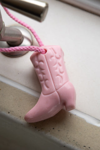 Image of the Pink Cowboy Boot Soap On A Rope hanging on a tap on a white kitchen surface.