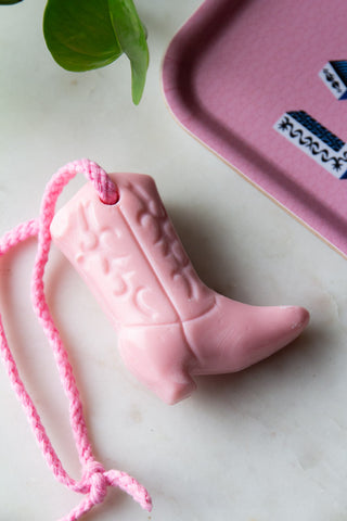 Close-up of the Pink Cowboy Boot Soap On A Rope styled on a white surface next to a pink tray and a plant.