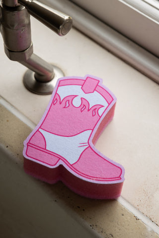 Image of the Pink Cowboy Boot Sponge styled next to a tap on a white kitchen surface.