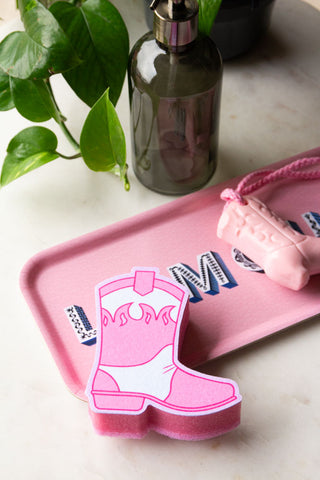 The Pink Cowboy Boot Sponge styled on a pink tray with a cowboy boot soap, next to a plant and a soap dispenser.