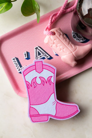 The Pink Cowboy Boot Sponge styled on a pink tray with a cowboy boot soap and a soap dispenser, next to a plant.