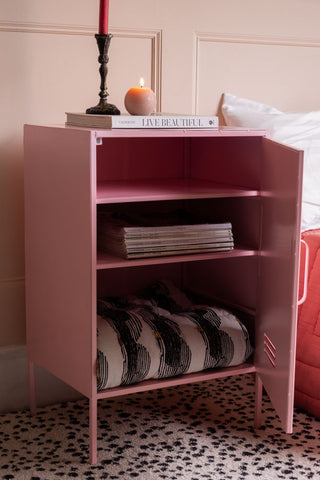 The Pink Industrial-Style Bedside Table with the door open, with magazines and a throw inside and various accessories on top, styled next to a bed.