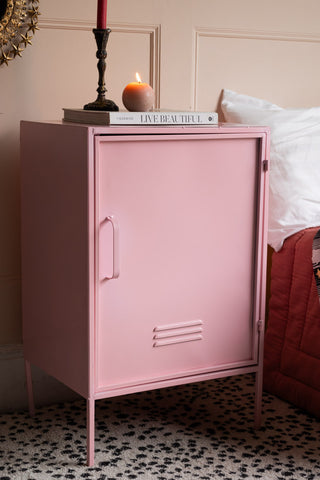 The Pink Industrial-Style Bedside Table styled with a lit candle, candlestick holder and book on, next to a bed.