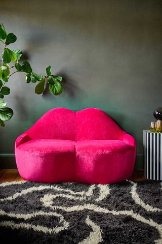 The Pink Lips Sofa styled with a rug, plant and side table with lamp and a glass.