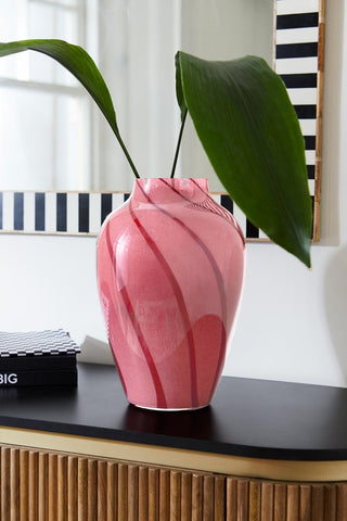 The Beautiful Pink & Red Swirl Vase styled on a wooden sideboard, with two large leaves inside.