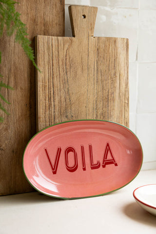 The Pink Voila Plate styled leaning against some wooden chopping boards in a kitchen.