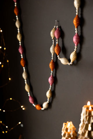 The Pink & Gold Sparkle Bauble Garland displayed draped on the wall, with some fairy lights and lit candles also in the shot.