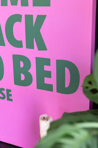 Close-up of the Pink & Green Come Back To Bed Please Canvas, with a candle and plant also in shot.