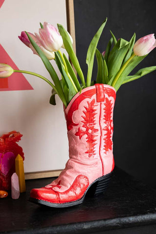 The Pink & Red Cowboy Boot Vase with flowers inside, on a black surface with colourful accessories.