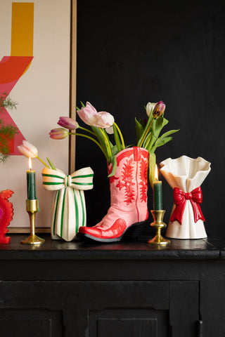 The Pink & Red Cowboy Boot Vase arranged on a surface with other vases, lit candles, a large art print and some flowers/greenery.