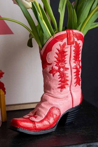 The Pink & Red Cowboy Boot Vase on a black surface with some flowers inside.