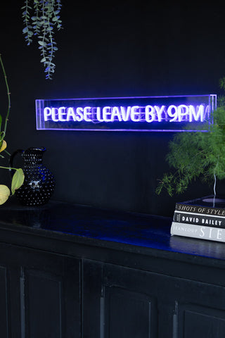 The Please Leave By 9pm Neon Lightbox illuminated and styled on a black wall above a sideboard, with plants, books and a jug.