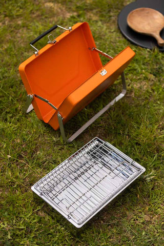 The Portable Suitcase BBQ - Burnt Orange in its separate parts, displayed on some grass.