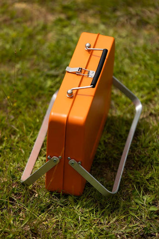 The Portable Suitcase BBQ - Burnt Orange displayed on some grass, leaning on its silver legs.