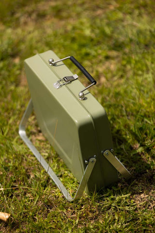 The Portable Suitcase BBQ - Khaki Green leaning on silver legs on some grass.