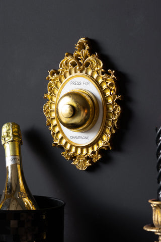 The Press For Champagne Wall Ornament displayed on a black wall behind an ice bucket with a gold bottle inside and a candle in a candlestick holder.