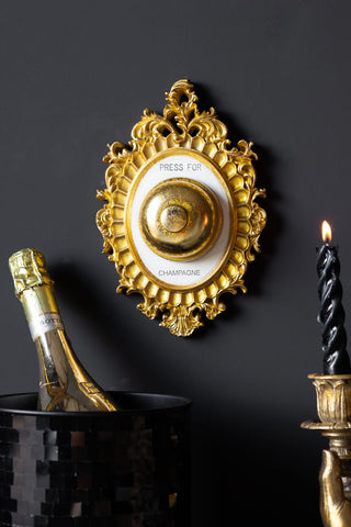 The Press For Champagne Wall Ornament displayed on a black wall, behind an ice bucket with a gold bottle of champagne inside and a lit candle in a candlestick holder.