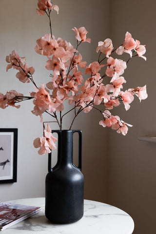 The Pretty Pink Blossom Flower Stem in a large black vase, displayed on a table with some magazines.