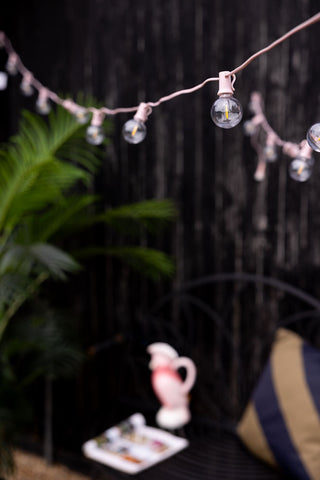 The Pretty Pink Festoon Lights  hanging in a garden, styled with various accessories and a plant.