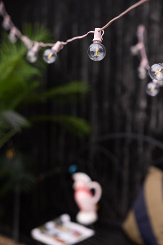 The Pretty Pink Festoon Lights hanging across a garden, styled with accessories and a plant.