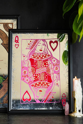 The Queen Of Hearts Decorative Playing Card Mirror styled on a black sideboard with another mirror, a crystal, a lit candle in a gold candlestick holder and some greenery.