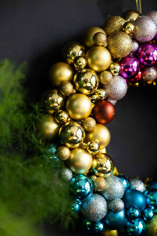 Close-up of the Rainbow Christmas Wreath hanging on a dark wall, styled above some greenery.