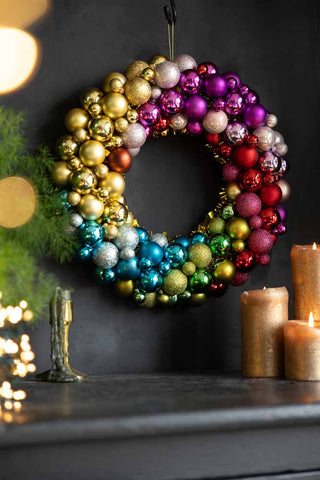 The Rainbow Christmas Wreath hanging on a dark wall above a sideboard styled with candles, fairy lights and greenery.