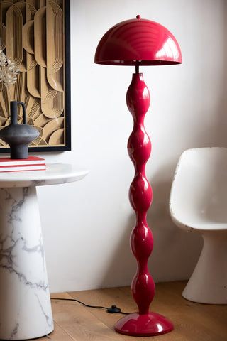 The Raspberry Red Wavy Retro Floor Light styled on a wooden floor next to a chair and a side table, styled with a book, vase with a dried flower in and a framed artwork on the wall in the background.