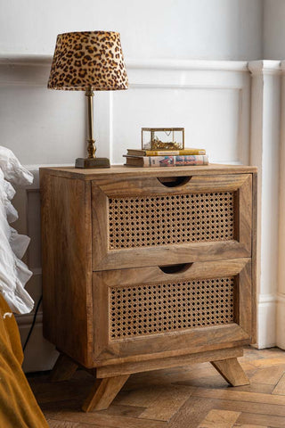 A two-drawer rattan bedside table, styled with a leopard print lamp, some books and a small box, styled next to a bed in a bedroom.