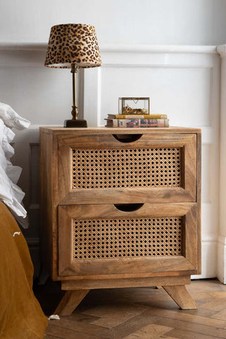 A rattan bedside table with two drawers, styled next to a bed with a leopard print lamp, some books and a small box.