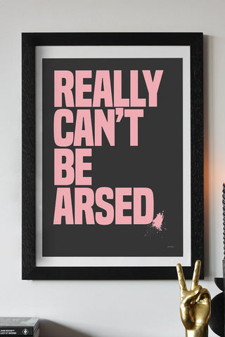 The Really Can't Be Arsed Black & Pink Art Print - Unframed styled in a black frame on a neutral wall above a hand ornament, books and candlestick holder.