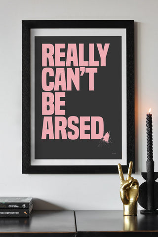 The Really Can't Be Arsed Black & Pink Art Print - Unframed styled in a black frame on a white wall, above a black surface with two books, a gold hand ornament and a lit candle in a candlestick holder.